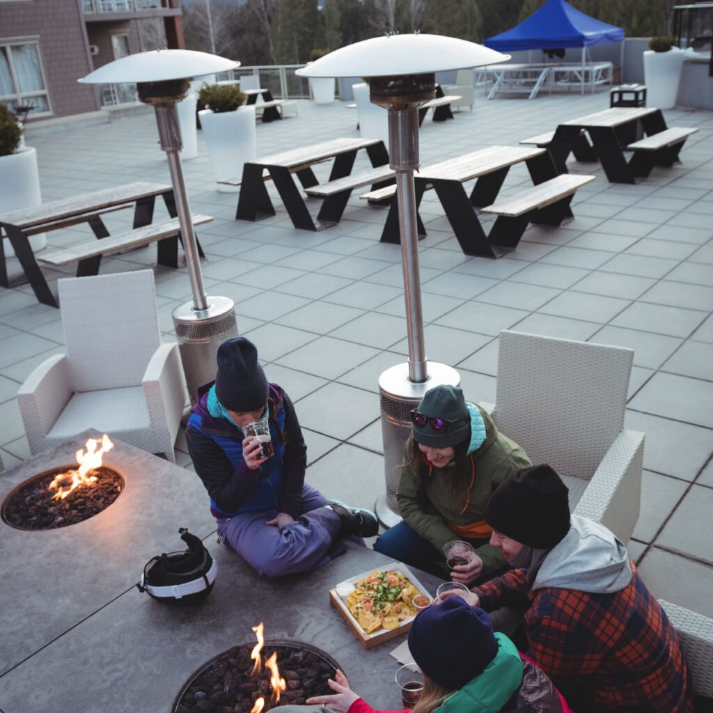 A group of travelers sitting around a fire pit at a hostel's outdoor lounge, sharing food and drinks with a scenic mountain backdrop. A warm and inviting atmosphere, showcasing the social aspect of solo travel